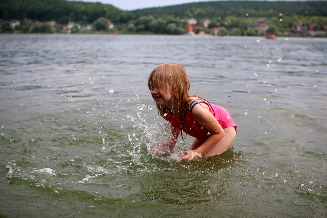 Smiling child splashing in the lake, enjoying a sunny day outdoors in a pink swimsuit