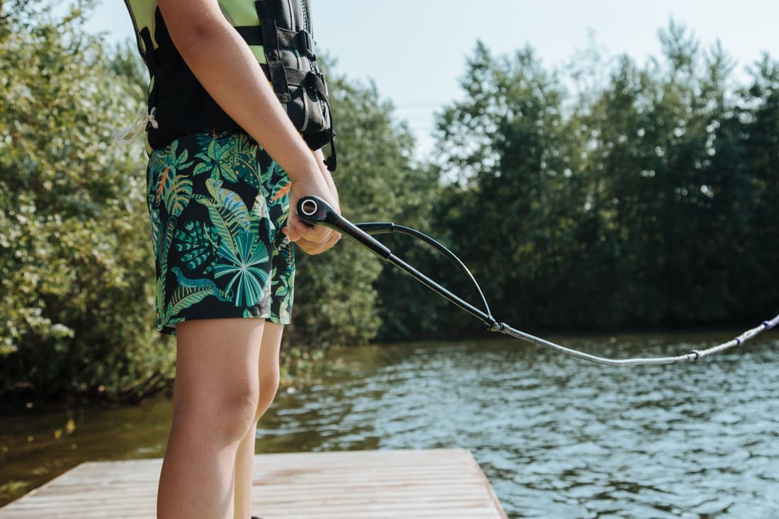 A child in life jacket prepares for a wakeboarding adventure on a sunny lakeside jetty