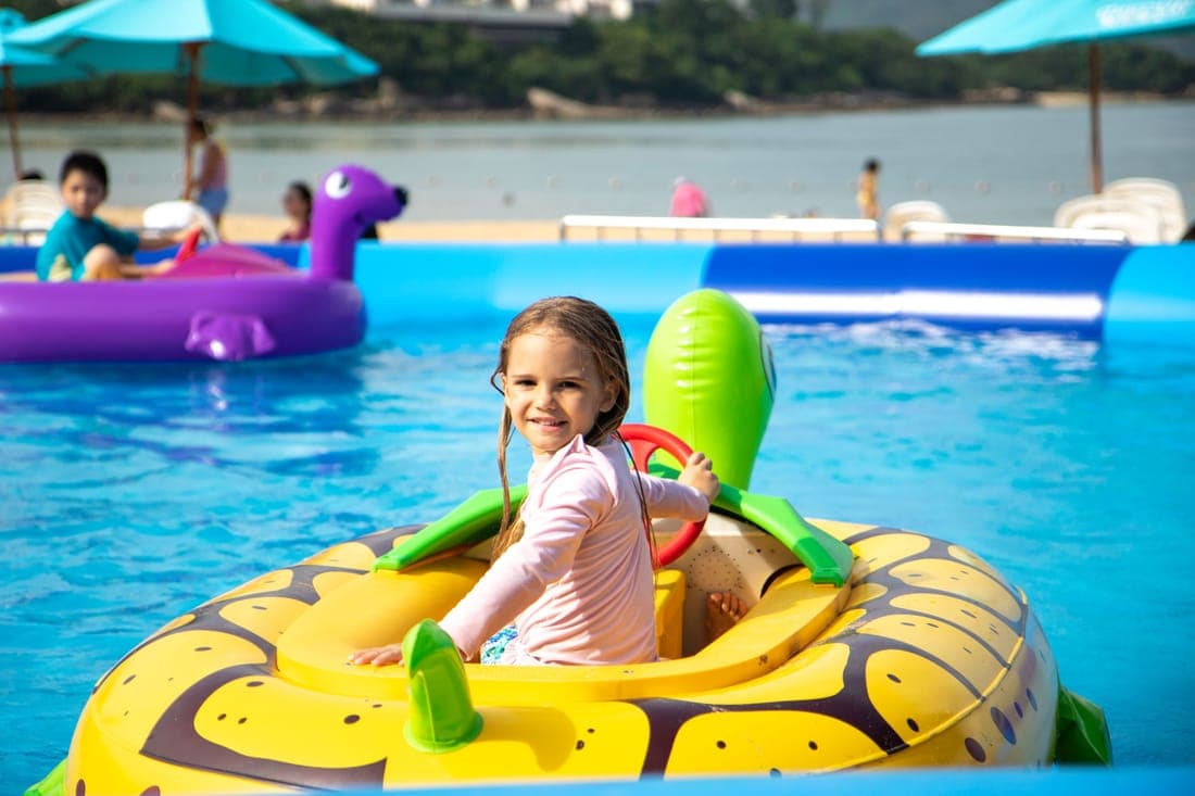 Happy child in pink sleeves enjoying a ride in a pool on a sunny day with inflatable toys around