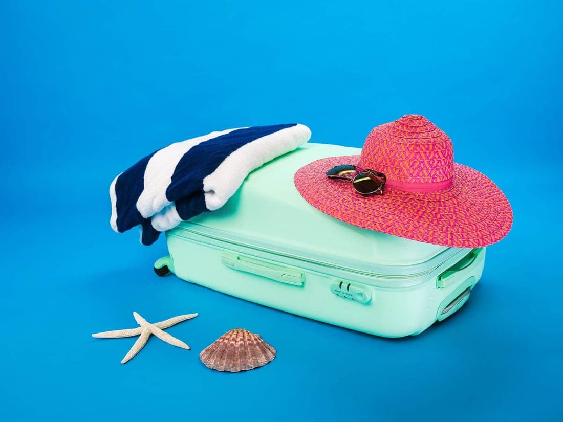 Beach essentials including a suitcase, hat, towel, and seashells on a bright background