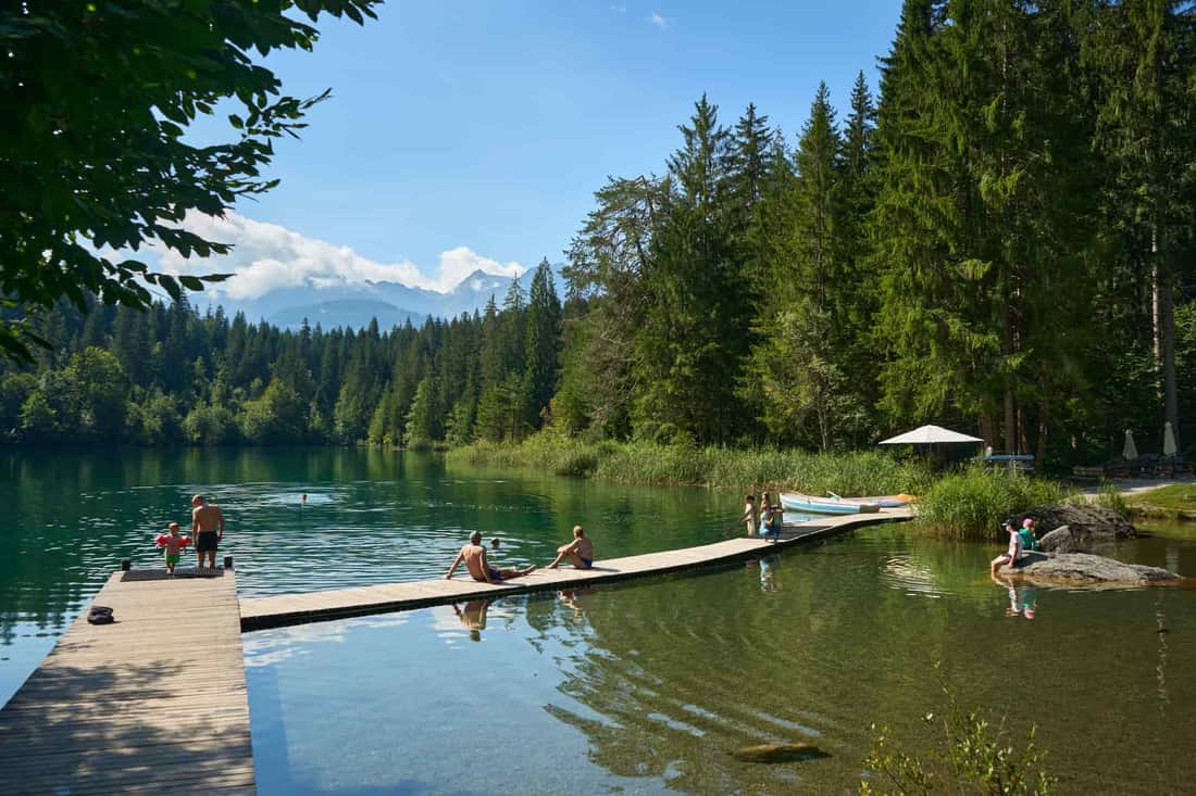 A serene lakeside view with people relaxing on a wooden dock surrounded by lush trees