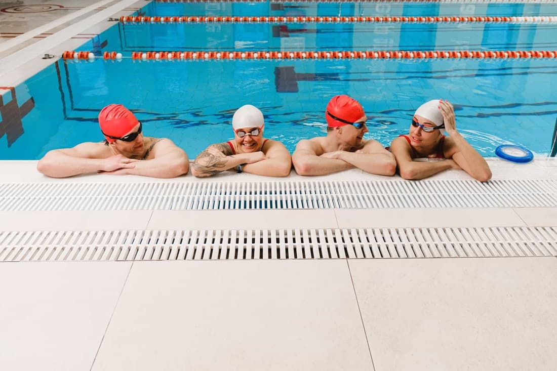 Four adult swimmers in caps and goggles relax by the indoor poolside, smiling and chatting