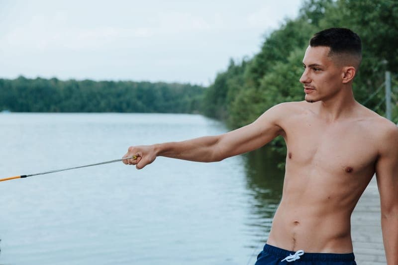 A shirtless man with a mustache and beard wakeboarding on a serene lake in summer