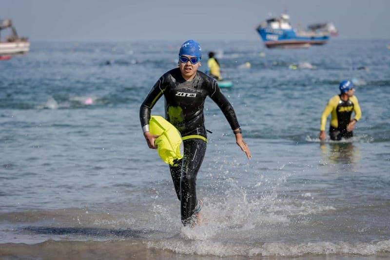 An athlete in a triathlon wetsuit emerges from the ocean, holding a swim buoy, during a competitive race