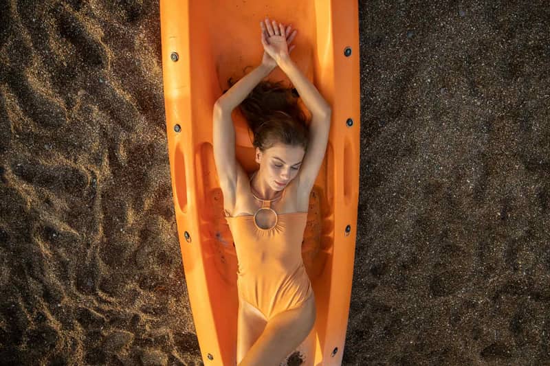 A woman in an orange one-piece relaxes on a kayak on a sandy beach